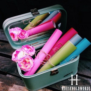 May include: A vintage green suitcase with a mirror inside, filled with colorful rolls of vinyl. The suitcase has a metal latch and a stitched design. The vinyl rolls are pink, red, green, blue, and gray. The suitcase is sitting on a wooden surface.