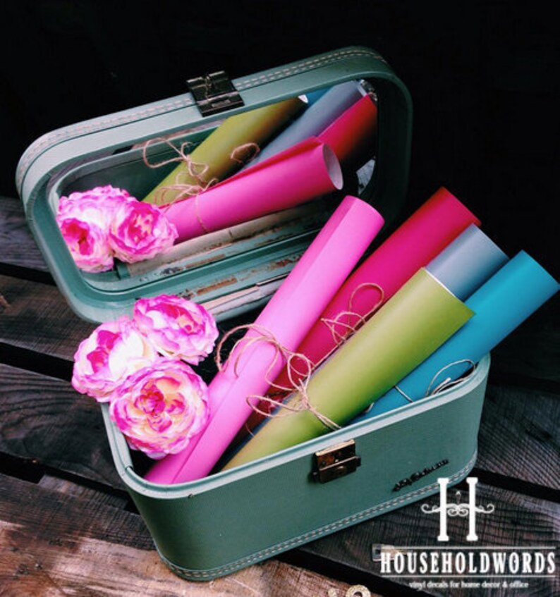 May include: A vintage green suitcase with a mirror inside, filled with colorful rolls of vinyl. The suitcase has a metal latch and a stitched design. The vinyl rolls are pink, red, green, blue, and gray. The suitcase is sitting on a wooden surface.
