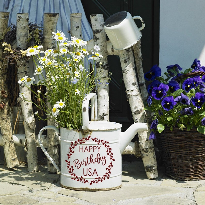 May include: White watering can with a red wreath and the text "Happy Birthday USA" on the side. The watering can is sitting on a stone patio with white birch branches and a pot of purple pansies in the background.