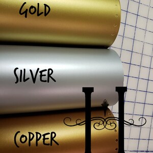 May include: Three rolls of metallic vinyl in gold, silver, and copper. The rolls are labelled with the colour names. The rolls are on a grid background.