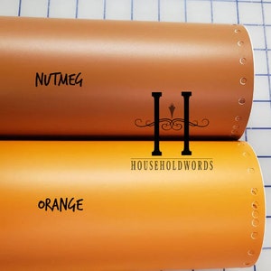 May include: Two rolls of vinyl adhesive film, one in a light brown colour labelled "NUTMEG" and the other in a bright orange colour labelled "ORANGE". The rolls are stacked on top of each other and have a white background.