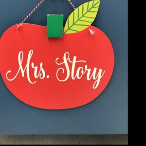 May include: Red apple-shaped sign with a green leaf and a white inscription that reads "Mrs. Story".