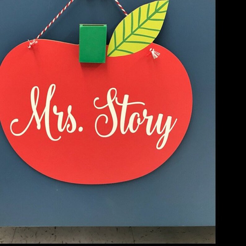 May include: Red apple-shaped sign with a green leaf and a white inscription that reads "Mrs. Story".