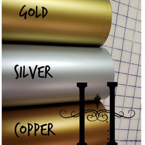 May include: Three rolls of metallic vinyl in gold, silver, and copper. The word "GOLD" is written in black on the gold roll, "SILVER" on the silver roll, and "COPPER" on the copper roll. The image also includes the text "HOUSEHOLDWORDS".