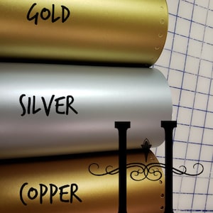 May include: Three rolls of metallic vinyl in gold, silver, and copper. The rolls are labeled with the color names. The rolls are on a grid background.