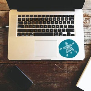 May include: A laptop computer with a blue and white sand dollar sticker on the top right corner of the trackpad. The laptop is on a wooden table with a notebook, pen, and a cup of coffee.