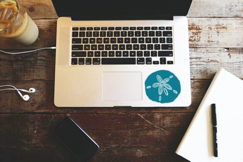 May include: A laptop computer with a blue and white sand dollar sticker on the top right corner of the trackpad. The laptop is on a wooden table with a notebook, pen, and a cup of coffee.