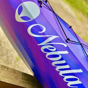 May include: A purple and blue kayak with a white alien logo and the word "Nebula" written in white cursive on the side.