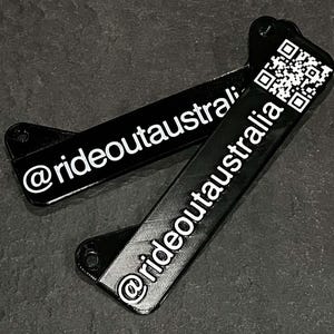 May include: Two black rectangular metal tags with rounded corners. The top tag has a QR code and the text "@rideoutaustralia". The bottom tag has the text "@rideoutaustralia".
