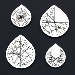 May include: Four white ceramic teardrop-shaped wall decorations with black thread embroidery designs. Each design is unique, featuring a different pattern of intersecting lines.