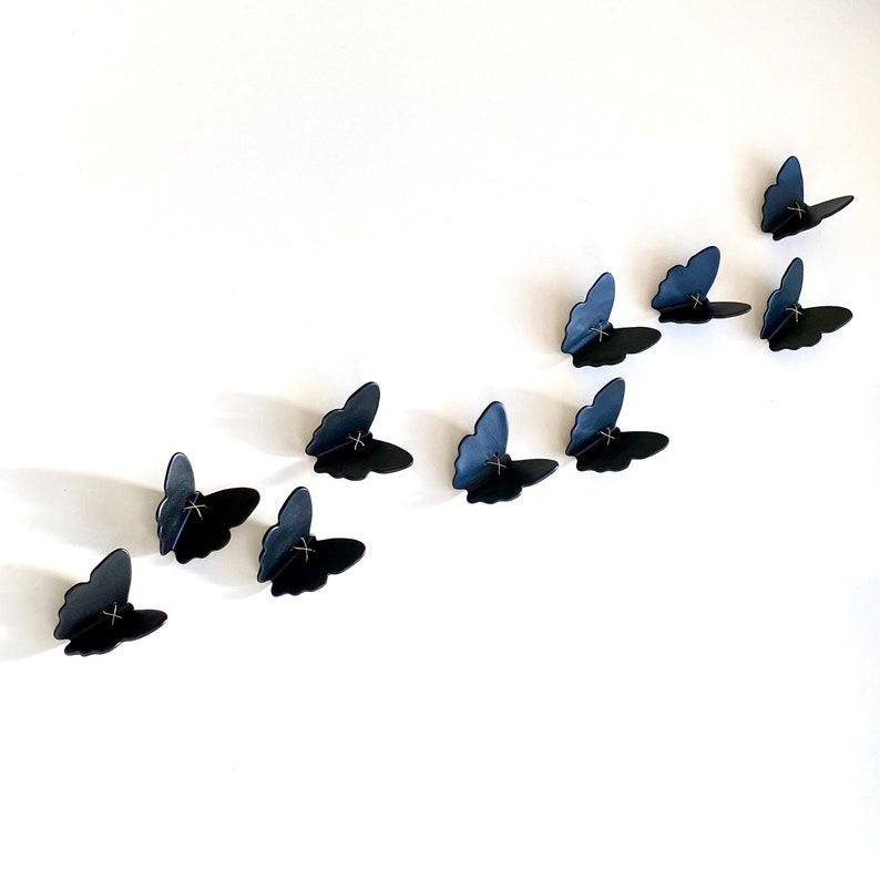 May include: A collection of 11 black butterfly wall decor pieces arranged in a diagonal line on a white wall.