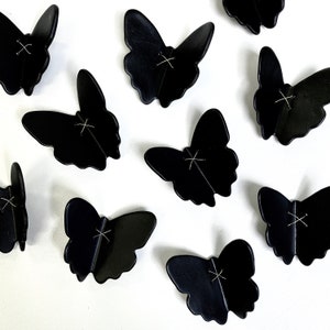 May include: Ten black butterfly-shaped ornaments with a stitched X design. The ornaments are arranged in a scattered pattern on a white background.