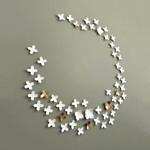 Oversized wall art comprising 60 ceramic sculptures in white and gold. Sculptural wall art, modern decor, to add texture to a wall. Each piece is roughly x shaped, like ultra simplified geometric flowers arranged like a wreath or garland on the wall.