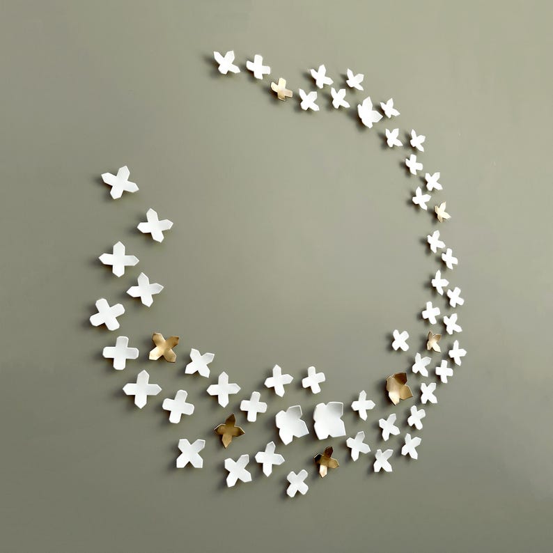 Oversized wall art comprising 60 ceramic sculptures in white and gold. Sculptural wall art, modern decor, to add texture to a wall. Each piece is roughly x shaped, like ultra simplified geometric flowers arranged like a wreath or garland on the wall.
