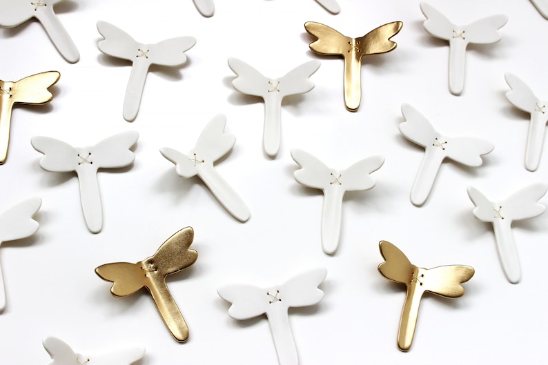 Original artwork, a close up of porcelain ceramic dragonfly sculptures. Minimalist design white and gold wall art dragonflies. Designed to flutter along your interior walls.
