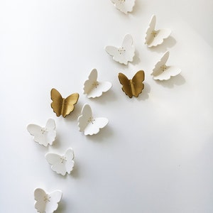 May include: A collection of white and gold ceramic butterflies, arranged in a diagonal pattern on a white wall. The butterflies are all different sizes and shapes, and some have a subtle texture.