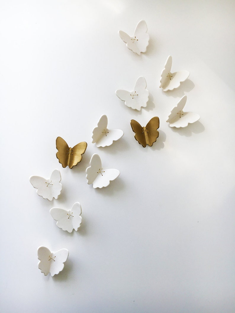 May include: A collection of white and gold ceramic butterflies, arranged in a diagonal pattern on a white wall. The butterflies are all different sizes and shapes, and some have a subtle texture.