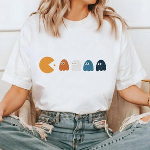May include: White t-shirt featuring a Pac-Man graphic and four ghosts in various colours. The ghosts are positioned in a line behind Pac-Man, who is consuming the first ghost.
