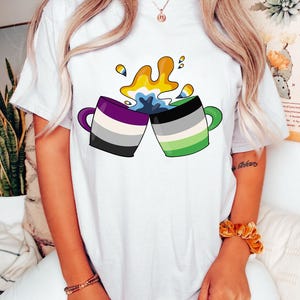 May include: White t-shirt with a graphic of two mugs clinking together, spilling colorful liquid. The mugs are decorated with the colors of the ace and genderqueer flags.