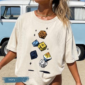 May include: An ivory short-sleeved t-shirt featuring a graphic print of five multi-sided gaming dice in orange, blue, yellow, dark blue, and white. The dice are illustrated with black outlines and numbers, surrounded by small black star symbols.