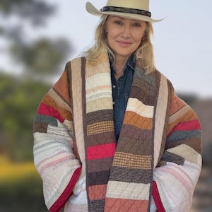 Puede incluir: Una chaqueta acolchada con un diseño de patchwork de rayas horizontales en tonos marrón, crema, rojo y gris. La prenda tiene mangas anchas, cuello tipo chal y un borde de color rojo oscuro, combinada con un sombrero de vaquero de color claro.