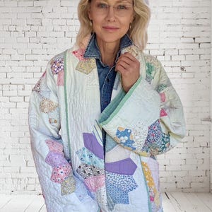 Könnte beinhalten: Gesteppte Jacke mit Patchwork-Design in Pastellfarben. Die Jacke hat eine Vielzahl von Blumen- und geometrischen Mustern mit hellgrünen Verzierungen. Die Jacke wird über einem Jeanshemd getragen.