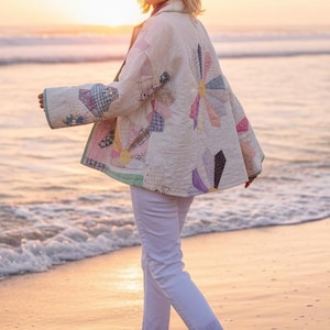 May include: A woman wearing a handmade, quilted jacket with a floral design, walking on a beach at sunset. The jacket features a variety of colorful fabric patches. The woman is also wearing white pants.