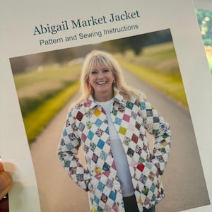 Op de afbeelding: Een gedrukt document getiteld Abigail Market Jacket Pattern and Sewing Instructions met een foto van een doorgestikt jasje met een witte achtergrond en een veelkleurig ruitpatroon, gedragen over een effen wit overhemd met kraag en zakken.