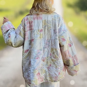 Puede incluir: Una chaqueta acolchada de manga larga con un diseño de patchwork que presenta varios patrones florales y de rayas en tonos pastel suaves de rosa, amarillo y verde. La chaqueta se combina con pantalones blancos y un sombrero de ala ancha.
