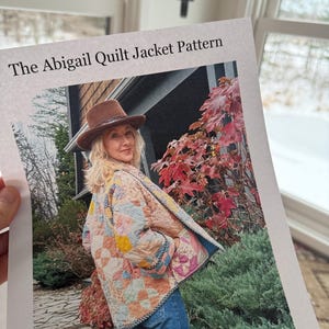 Może przedstawiać: Wydrukowany wzór zatytułowany "The Abigail Quilt Jacket Pattern" jest trzymany przed oknem. Wzór przedstawia zdjęcie kobiety w kolorowej kurtce z patchworku, niebieskich dżinsach i brązowym kapeluszu. Kurtka ma kwiatowy wzór.