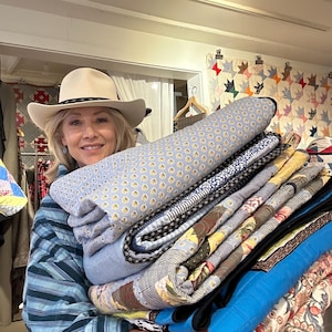 May include: A woman wearing a white cowboy hat and a blue and white striped shirt is holding a stack of colorful quilts. The quilts are folded and stacked on top of each other. The woman is smiling and looking at the camera.