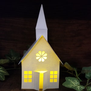 May include: A white paper church with a lit window and a flower design on the front. The church is sitting on a wooden surface with green leaves.