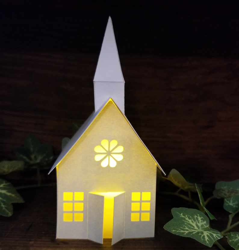 May include: A white paper church with a lit window and a flower design on the front. The church is sitting on a wooden surface with green leaves.