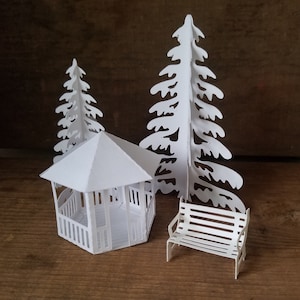 May include: White papercraft model of a gazebo with a white bench and two white trees. The gazebo has a white roof and a white railing. The bench has a white slatted design. The trees have a white silhouette design.