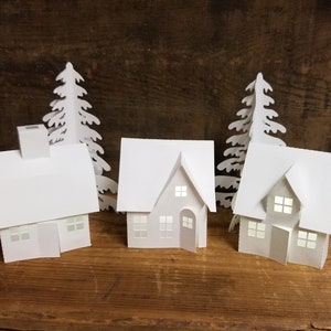 May include: Three white paper houses with windows and doors, and two white paper trees, all on a wooden surface.