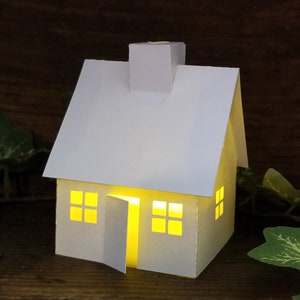 May include: A white paper house with a chimney and windows. The house is lit from inside, creating a warm glow.