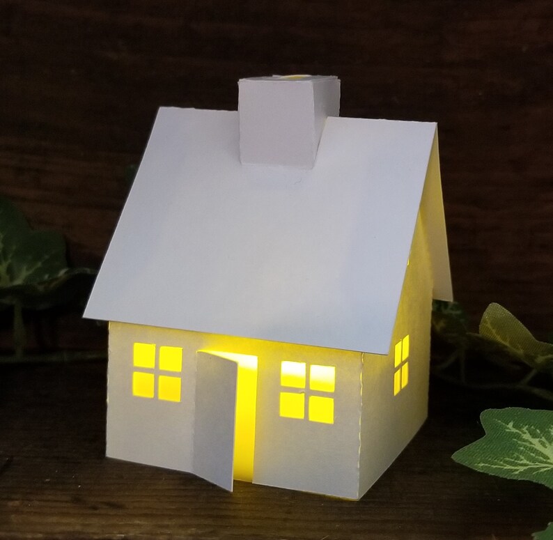May include: A white paper house with a chimney and windows. The house is lit from inside, creating a warm glow.