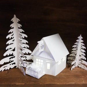May include: White papercraft model of a small house with a porch and two white paper trees. The house has a white roof and a white door. The trees are cut out of paper and have a simple design.