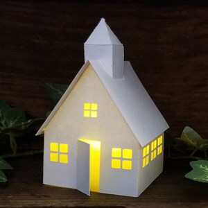 May include: A white paper model of a house with a lit window and open door. The house has a pointed roof and a small steeple. The house is sitting on a wooden surface.