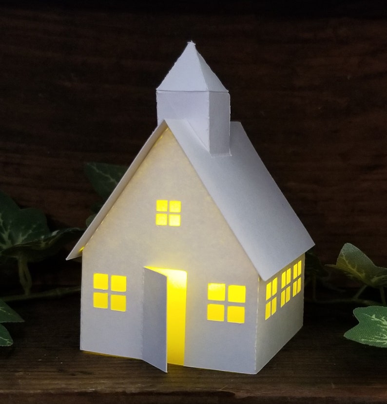 May include: A white paper model of a house with a lit window and open door. The house has a pointed roof and a small steeple. The house is sitting on a wooden surface.