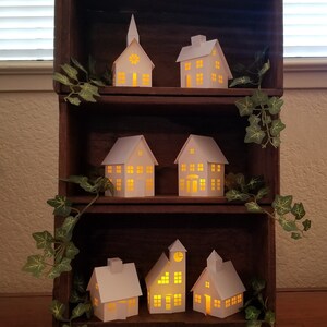 May include: A wooden shelf unit with six white paper houses with lit windows. The houses are arranged in two rows of three. The shelf unit is decorated with green ivy.