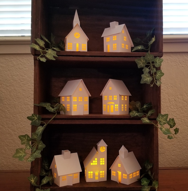May include: A wooden shelf unit with six white paper houses with lit windows. The houses are arranged in two rows of three. The shelf unit is decorated with green ivy.