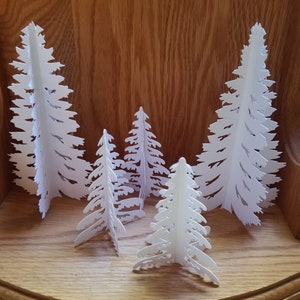May include: Five white paper Christmas trees of varying sizes, standing on a wooden surface. The trees are cut out in a delicate, intricate design.