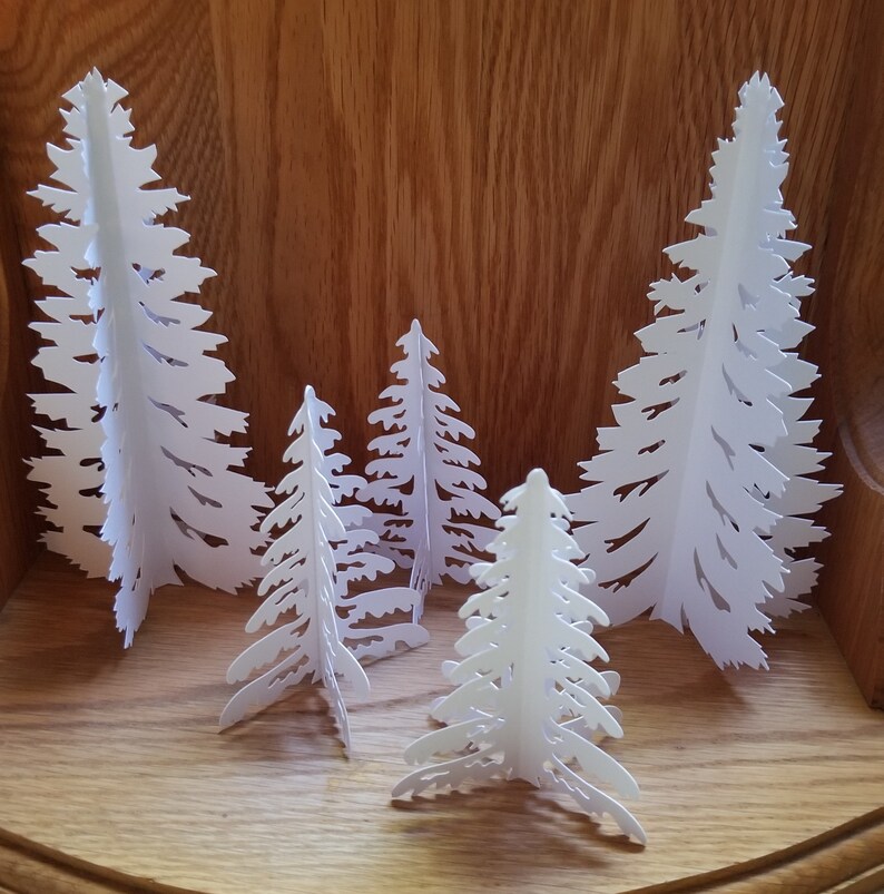 May include: Five white paper Christmas trees of varying sizes, standing on a wooden surface. The trees are cut out in a delicate, intricate design.