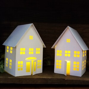 May include: Two white paper houses with cut-out windows and doors, illuminated from within with warm yellow light. The houses are sitting on a wooden surface.