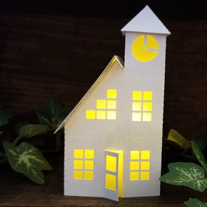 May include: A white paper model of a house with a clock tower. The house has windows and a door, all of which are lit from within. The house is sitting on a wooden surface.