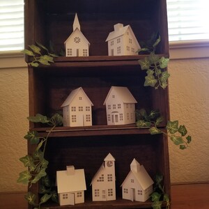 May include: A wooden shelf unit with three shelves, each displaying a collection of white paper model houses. The houses are in various styles, including a church, a cottage, and a schoolhouse. The shelf unit is decorated with green ivy.