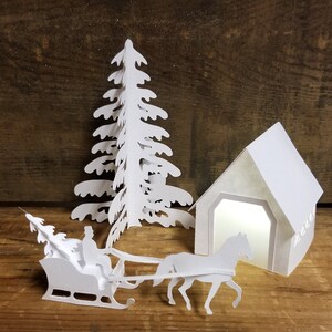 May include: White papercraft scene with a white paper house, a white paper tree, and a white paper horse-drawn sleigh. The house has a window with a light shining through it. The sleigh is pulled by a horse and has a person sitting in the front.