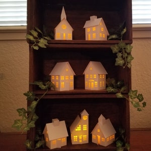 May include: A wooden shelf unit with three shelves, each displaying a collection of white paper houses with glowing windows. The houses are arranged in a staggered pattern, creating a charming and festive display. The shelf unit is decorated with green ivy vines.