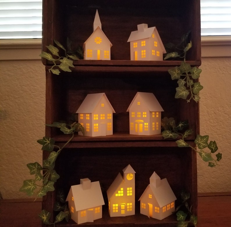 May include: A wooden shelf unit with three shelves, each displaying a collection of white paper houses with glowing windows. The houses are arranged in a staggered pattern, creating a charming and festive display. The shelf unit is decorated with green ivy vines.